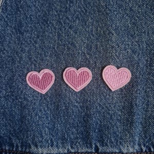 May include: Three small, light pink heart-shaped embroidered patches are arranged in a horizontal row. Each patch features a solid pink centre with a lighter pink stitched border. The patches are attached to a dark blue denim fabric background.