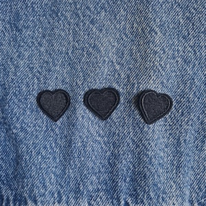 May include: A medium-wash blue denim fabric featuring three small, dark navy blue embroidered heart-shaped patches arranged in a horizontal row across the centre of the material, showcasing a classic twill texture and visible stitching.