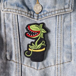 May include: An embroidered patch featuring a green carnivorous plant with a red mouth and white teeth, growing out of a black pot with a yellow rim. The patch is attached to a light blue denim jacket with a silver metal button visible above it.