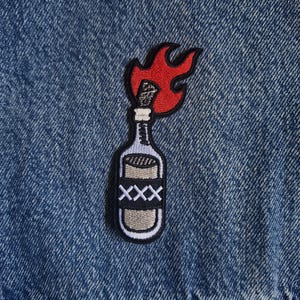 May include: An embroidered patch featuring a glass bottle with a lit fuse and red flames emerging from the top. The bottle has a black label with three white X symbols and is attached to a blue denim fabric background with visible stitching.