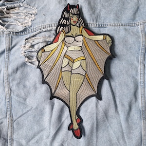 Op de afbeelding: Geborduurde patch met een pin-up-achtige vleermuisvrouw met zwart haar, rode hoorns en vleermuisvleugels. De patch heeft een lichtgrijs, geel en rood kleurenschema. De patch is bevestigd op een lichtblauwe spijkerjas.