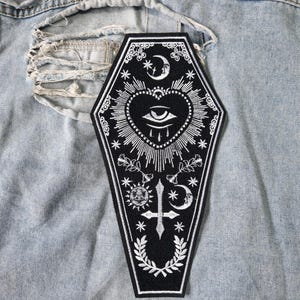 May include: A black coffin-shaped embroidered patch featuring white thread details of a central eye within a heart, a sun, a moon, stars, a cross, and floral elements. The patch is displayed against a light blue distressed denim fabric background.