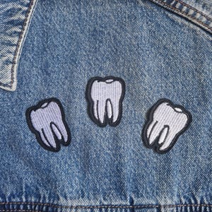 Peut inclure: Une veste en jean bleu ornée de trois écussons brodés en forme de dent. Les motifs sont bordés de fil noir, avec une dent violet clair à gauche, une dent lavande au centre et une dent blanche à droite, le tout cousu sur le tissu.