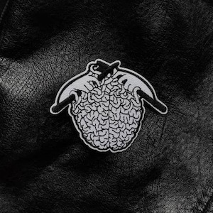Op de afbeelding: Een zwart leren jack met een wit geborduurd patch van een brein dat door twee handen wordt vastgehouden.
