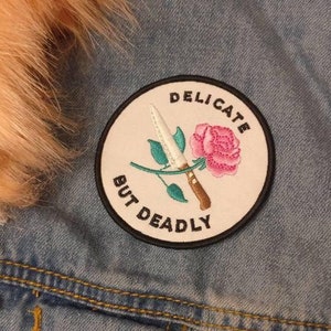Op de afbeelding: Een spijkerjack met een witte ronde patch waarop "Delicate But Deadly" staat, met een roze roos en een mes in het midden.