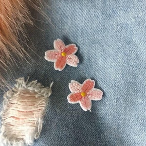 Puede incluir: Primer plano de dos parches bordados con flores rosas con centros amarillos sobre un fondo de mezclilla azul claro. Los parches tienen forma de flor de cerezo. También se ve un parche de tela deshilachada y piel rosa.
