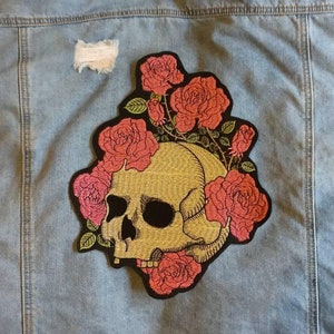 May include: A blue denim jacket with a skull and roses embroidered patch on the back. The patch is in shades of pink, yellow, and green.