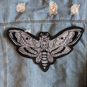 以下が含まれることがあります： 複雑なディテールが施された黒と白の蛾のパッチが、ダメージ加工が施されたライトブルーのデニムジャケットに縫い付けられています。