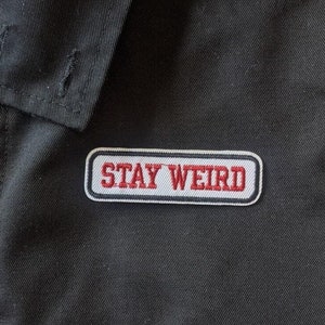 Peut inclure: Veste noire avec une doublure en tartan rouge et blanc et un patch gris sur lequel est inscrit "STAY WEIRD" en lettres rouges.