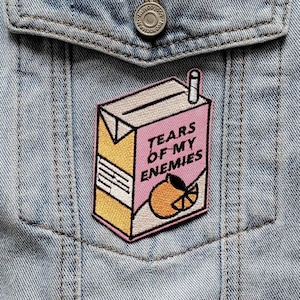 May include: Pale blue denim jacket featuring a patch on the pocket. The patch is a juice box design with the text "TEARS OF MY ENEMIES" in pink and a slice of orange. The jacket has distressed details.