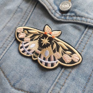 K&ouml;nnte beinhalten: Ein gestickter Motten-Patch in Schwarz und Gold mit rosa und wei&szlig;en Akzenten. Die Motte hat einen Stern auf ihrem K&ouml;rper und ist von floralen Details umgeben.