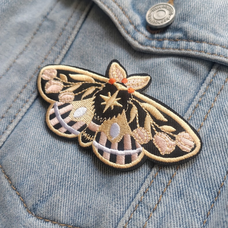 K&ouml;nnte beinhalten: Ein gestickter Motten-Patch in Schwarz und Gold mit rosa und wei&szlig;en Akzenten. Die Motte hat einen Stern auf ihrem K&ouml;rper und ist von floralen Details umgeben.