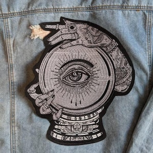 May include: A black and white embroidered patch depicting a hand holding a crystal ball with an eye inside. The hand is decorated with intricate designs.