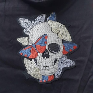 May include: Embroidered patch showing a skull design adorned with butterflies. The skull is white with detailed stitching, and the butterflies have red, blue, and cream-coloured wings. The patch is on a black background.