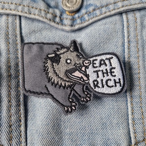 May include: Embroidered patch featuring a gray opossum with an open mouth and the text "EAT THE RICH" in a speech bubble. The patch is rectangular with a black border and is affixed to a denim jacket.