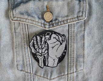 May include: Black and white embroidered patch of a skeleton hand and a human hand holding each other. The patch is circular and has a white outline.