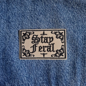 May include: A rectangular embroidered patch featuring a beige background with a black decorative border and the text Stay Feral in a gothic-style font. The patch is attached to a blue denim fabric with a visible twill texture and stitching.