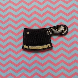 Op de afbeelding: Een geborduurde patch van een slagersmes, in zwart en goud.
