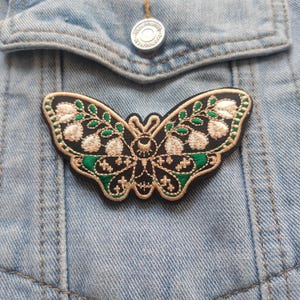 K&ouml;nnte beinhalten: Ein schwarzer gestickter Schmetterlings-Patch mit goldenen und gr&uuml;nen F&auml;den. Der Schmetterling hat einen Halbmond und Sterne auf seinem K&ouml;rper.
