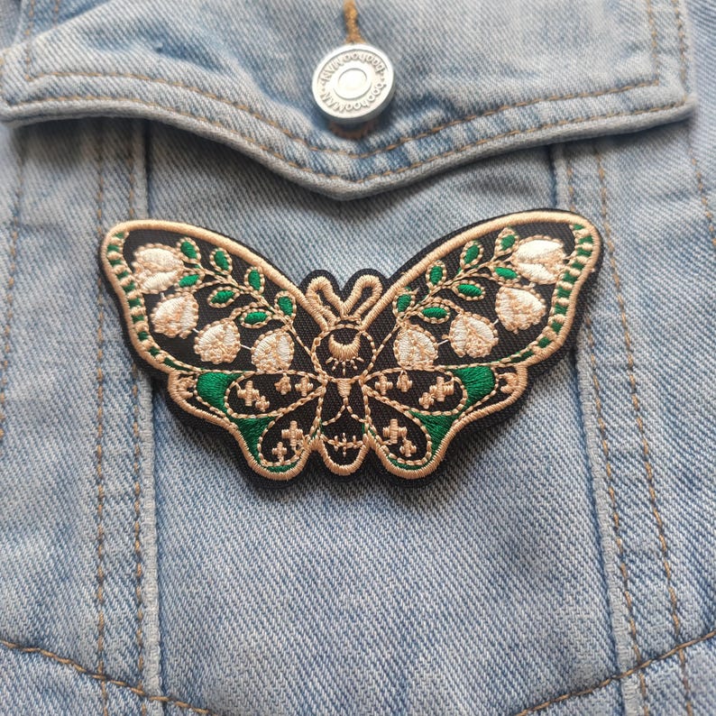K&ouml;nnte beinhalten: Ein schwarzer gestickter Schmetterlings-Patch mit goldenen und gr&uuml;nen F&auml;den. Der Schmetterling hat einen Halbmond und Sterne auf seinem K&ouml;rper.