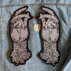 Könnte beinhalten: Zwei schwarze Bügeleisen-Patches mit einem silbernen gestickten Design von Händen mit Rosen. Die Hände sind in einer Gebetsposition mit ineinander verschlungenen Fingern. Die Rosen befinden sich am unteren Ende der Hände.