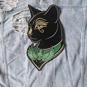May include: An embroidered patch featuring a black cat head with gold outlines and an Eye of Horus detail. The cat wears a green and gold collar adorned with a winged scarab beetle. The patch is placed on a light blue distressed denim fabric.