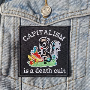 May include: A square black embroidered patch featuring a cartoon skunk sitting on green grass next to a red mushroom and a blue bird. The text CAPITALISM is arched above the skunk, and the text is a death cult is printed below in white.
