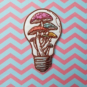 Op de afbeelding: Een geborduurde strijk-patch met een gloeilamp met kleurrijke paddenstoelen erin. De gloeilamp is wit met een bruin filament en een zilveren voet. De paddenstoelen zijn roze, blauw, oranje en geel.