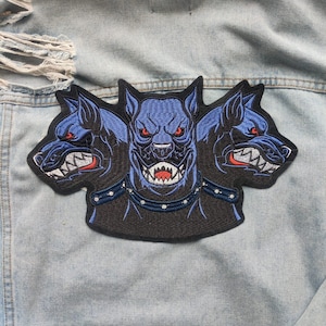May include: A black and blue embroidered patch of a three-headed dog with a studded collar. The dog has red eyes and sharp teeth.
