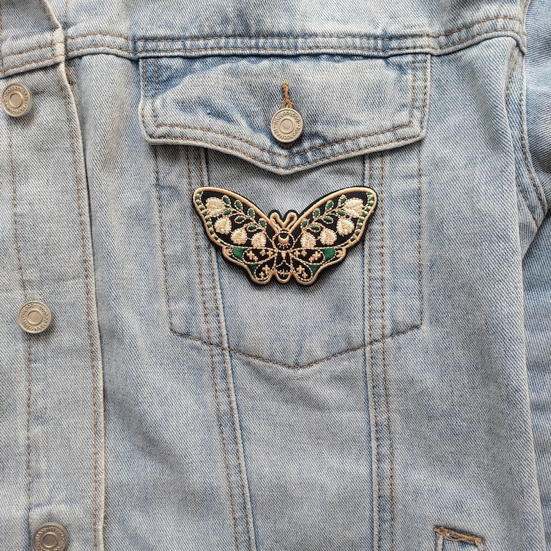 K&ouml;nnte beinhalten: Ein gestickter Schmetterlings-Patch in Schwarz und Gr&uuml;n mit goldenen Akzenten. Der Patch ist an einer Jeansjacke befestigt.