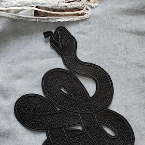 Op de afbeelding: Zwarte geborduurde slangenpatch op een lichtblauwe spijkerjas. De slang is opgerold en gedetailleerd, met een getextureerd uiterlijk. De denim heeft distressed plekken.