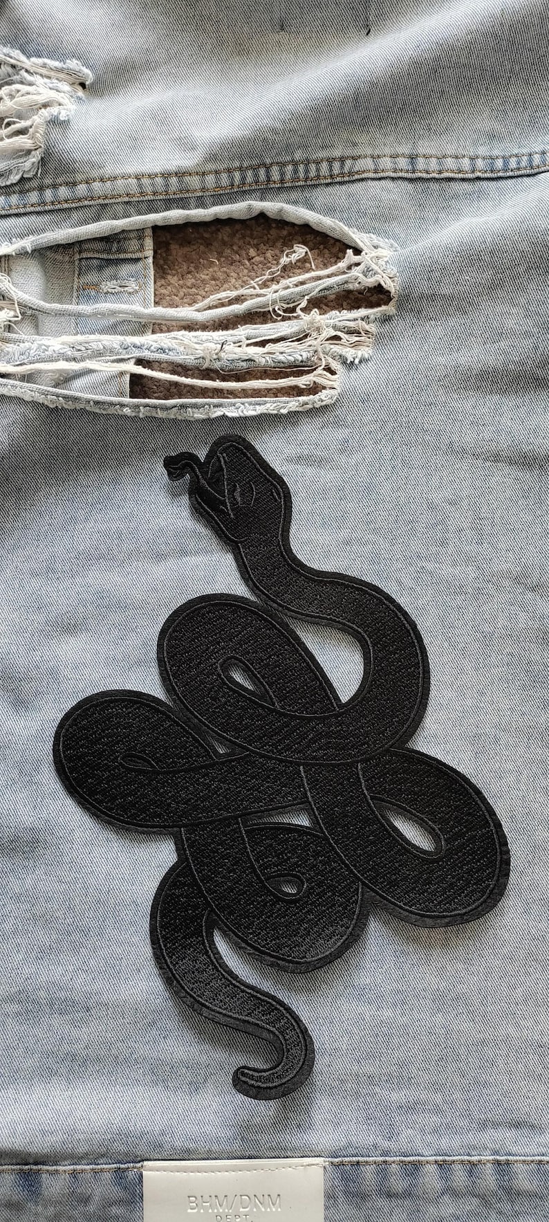 Op de afbeelding: Zwarte geborduurde slangenpatch op een lichtblauwe spijkerjas. De slang is opgerold en gedetailleerd, met een getextureerd uiterlijk. De denim heeft distressed plekken.