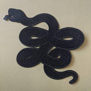 Op de afbeelding: Een zwart geborduurd embleem met een ontwerp van een opgerolde slang. De slang is afgebeeld met zijn tong uitgestoken en het embleem heeft een getextureerd uiterlijk. Het embleem is geplaatst tegen een lichtbeige achtergrond.