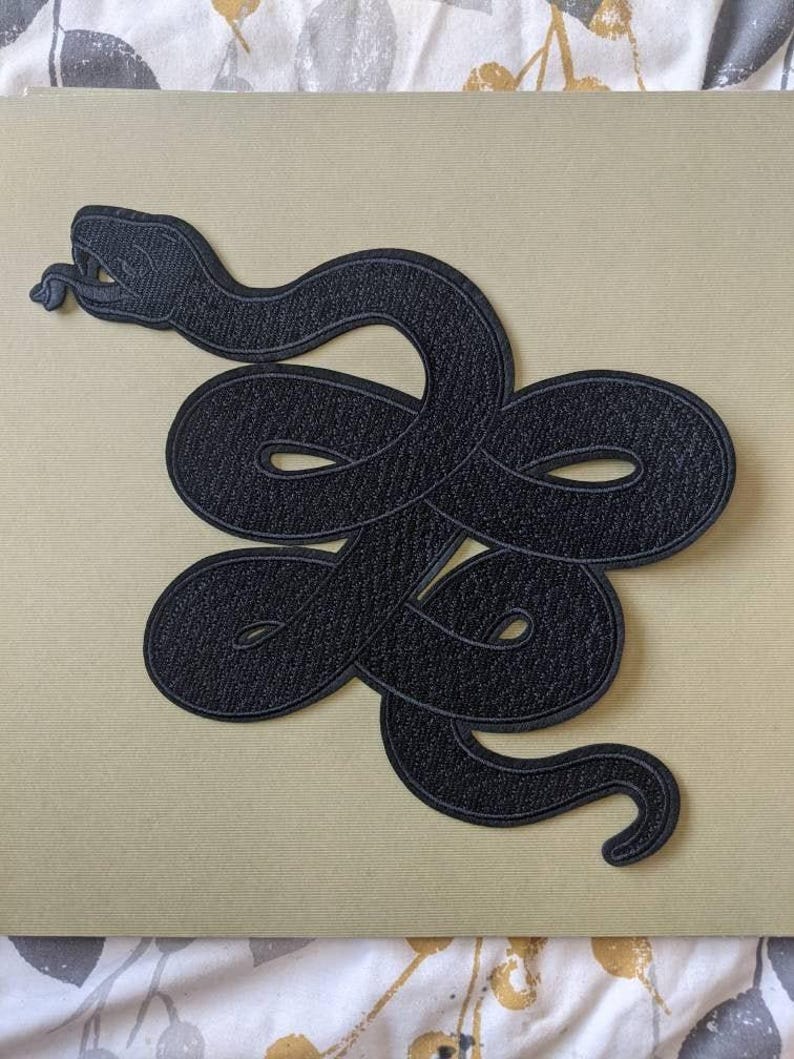 Op de afbeelding: Een zwart geborduurd embleem met een ontwerp van een opgerolde slang. De slang is afgebeeld met zijn tong uitgestoken en het embleem heeft een getextureerd uiterlijk. Het embleem is geplaatst tegen een lichtbeige achtergrond.