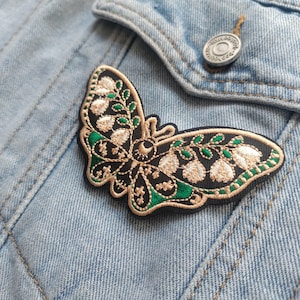 K&ouml;nnte beinhalten: Ein gestickter Schmetterlings-Patch in Schwarz und Gr&uuml;n mit goldenen Akzenten. Der Schmetterling hat ein detailliertes Design mit Bl&auml;ttern und einem mondf&ouml;rmigen Auge.