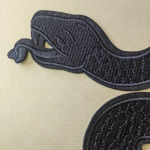 Op de afbeelding: Zwarte geborduurde slangenpatch met gedetailleerde stiksels. De patch toont een slangenkop en een opgerold lichaamssegment. Het ontwerp staat op een beige achtergrond. Geschikt voor jassen, tassen of andere stoffen items.