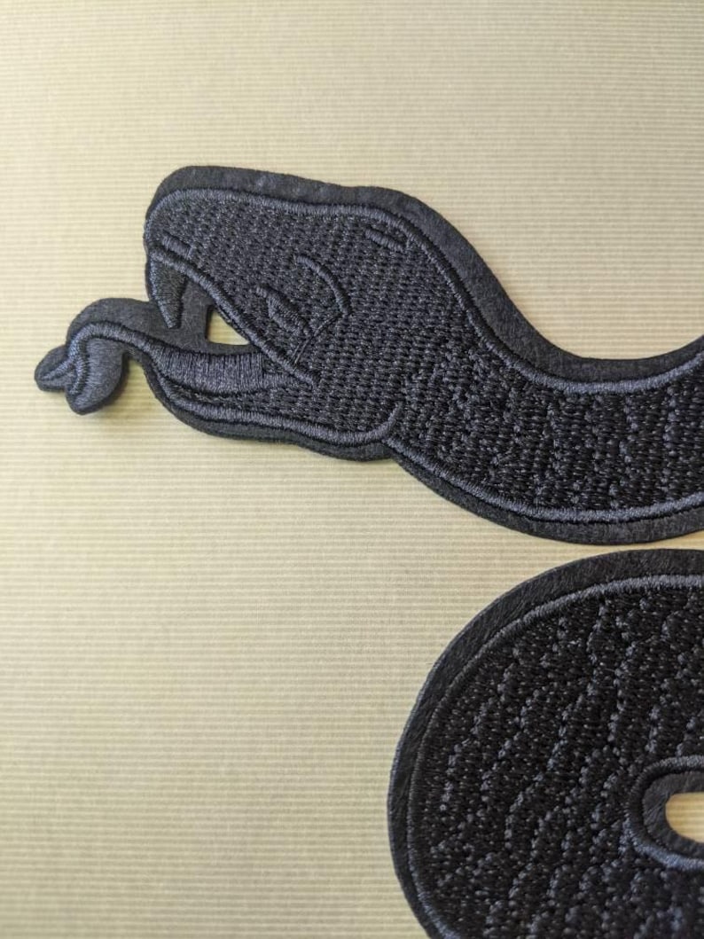 Op de afbeelding: Zwarte geborduurde slangenpatch met gedetailleerde stiksels. De patch toont een slangenkop en een opgerold lichaamssegment. Het ontwerp staat op een beige achtergrond. Geschikt voor jassen, tassen of andere stoffen items.