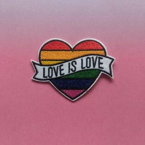 Peut inclure: Un patch en forme de cœur arc-en-ciel avec le texte "LOVE IS LOVE" sur une bannière blanche. Le cœur est bordé de noir et présente un motif de rayures arc-en-ciel.