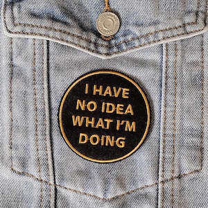 Puede incluir: Un parche bordado en negro y dorado cosido en una chaqueta vaquera azul. El parche dice "No tengo ni idea de lo que estoy haciendo."