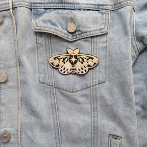K&ouml;nnte beinhalten: Ein gestickter Motten-Patch in Schwarz und Gold mit rosa und wei&szlig;en Blumendetails. Der Patch ist auf eine hellblaue Jeansjacke gen&auml;ht.