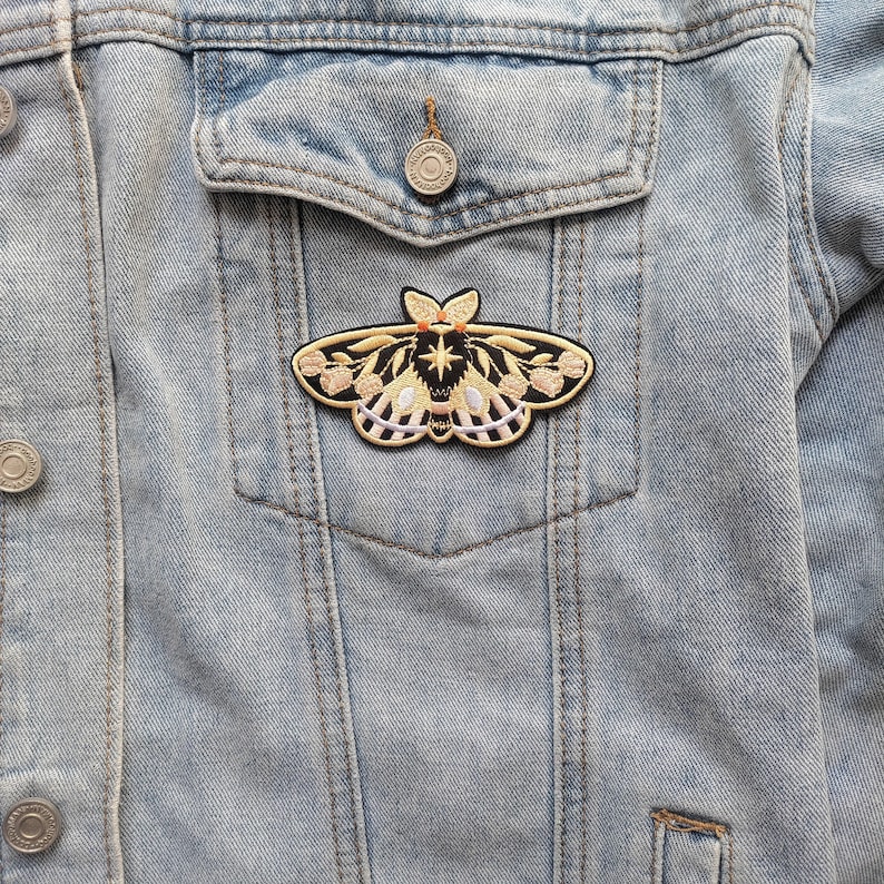K&ouml;nnte beinhalten: Ein gestickter Motten-Patch in Schwarz und Gold mit rosa und wei&szlig;en Blumendetails. Der Patch ist auf eine hellblaue Jeansjacke gen&auml;ht.