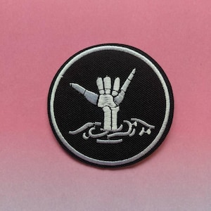 Peut inclure: Un patch brodé noir et blanc avec une main squelettique faisant le signe shaka, entouré d'un cercle blanc et de vagues noires. Le texte "SCREAM" est brodé en blanc sous la main.