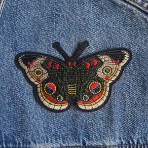 Könnte beinhalten: Eine blaue Jeansjacke mit einem aufgestickten Schmetterlingsaufnäher. Der Schmetterling hat dunkelgrüne Flügel mit kreisförmigen Mustern in Rot, Gelb und Blau, akzentuiert durch kleine rote Herzformen und einen weißen Rand an den Flügelunterkanten.