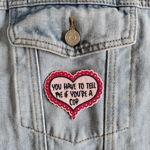 Könnte beinhalten: Rosa herzförmiger Patch mit dem Text "You have to tell me if you're a cop" auf einer Jeansjacke.