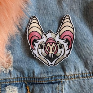 May include: An embroidered patch featuring the head of a bat with white, cream, and pink coloring, black outlines, and two small white teeth. The patch is attached to a blue denim fabric surface with a visible metal button and frayed edges.