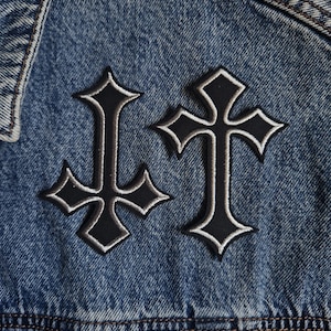 May include: Two embroidered patches featuring black crosses with silver-grey borders are attached to a blue denim jacket. One cross is oriented upright, while the other is inverted. The denim fabric shows a classic blue wash with visible texture.