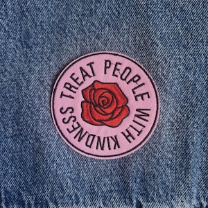 May include: A circular embroidered patch attached to blue denim fabric. The patch features a light pink background with a red rose in the center and the black text "TREAT PEOPLE WITH KINDNESS" arranged in a circle around the rose design.