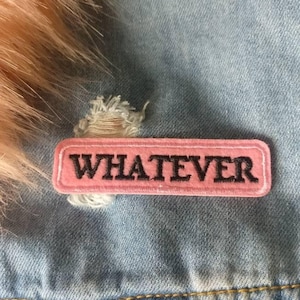 May include: A pink rectangular patch with the word "WHATEVER" in black lettering is sewn onto a light blue denim jacket. The patch is slightly distressed. Peach-coloured faux fur trim is visible at the top of the image.