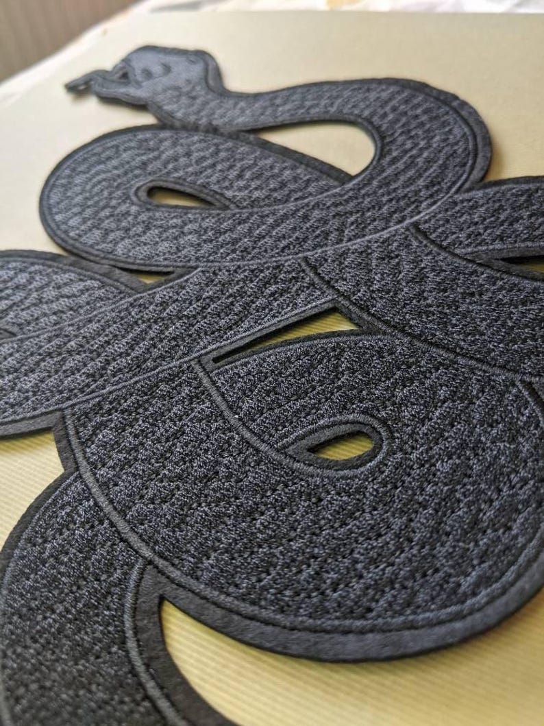 Op de afbeelding: Close-up van een donkergrijze geborduurde slangenpatch met een getextureerd oppervlak. De slang is opgerold en gedetailleerd, met een zwarte rand. De patch is waarschijnlijk voor kleding of accessoires.