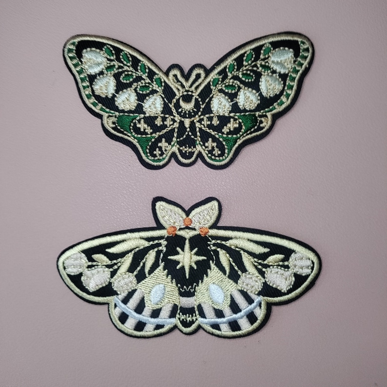 K&ouml;nnte beinhalten: Zwei gestickte B&uuml;gelbilder mit Mottenmotiven. Das obere Patch ist gr&uuml;n und gold mit einem schwarzen K&ouml;rper und das untere Patch ist schwarz und gold mit einem wei&szlig;-schwarz gestreiften K&ouml;rper.